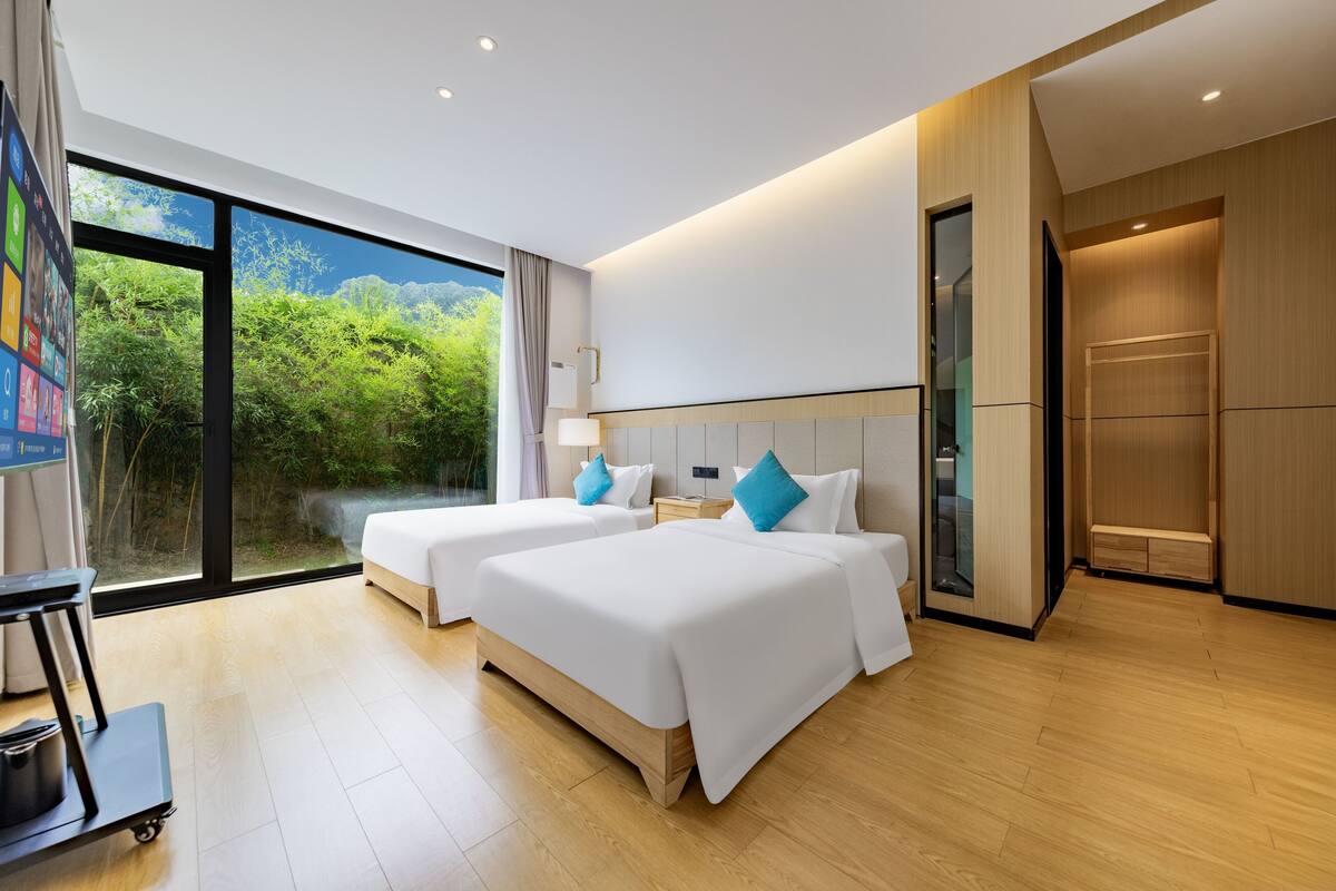 Une chambre d’hôtel moderne avec deux lits, une grande fenêtre donnant sur un espace verdoyant, une kitchenette et une porte coulissante menant à la salle de bain.