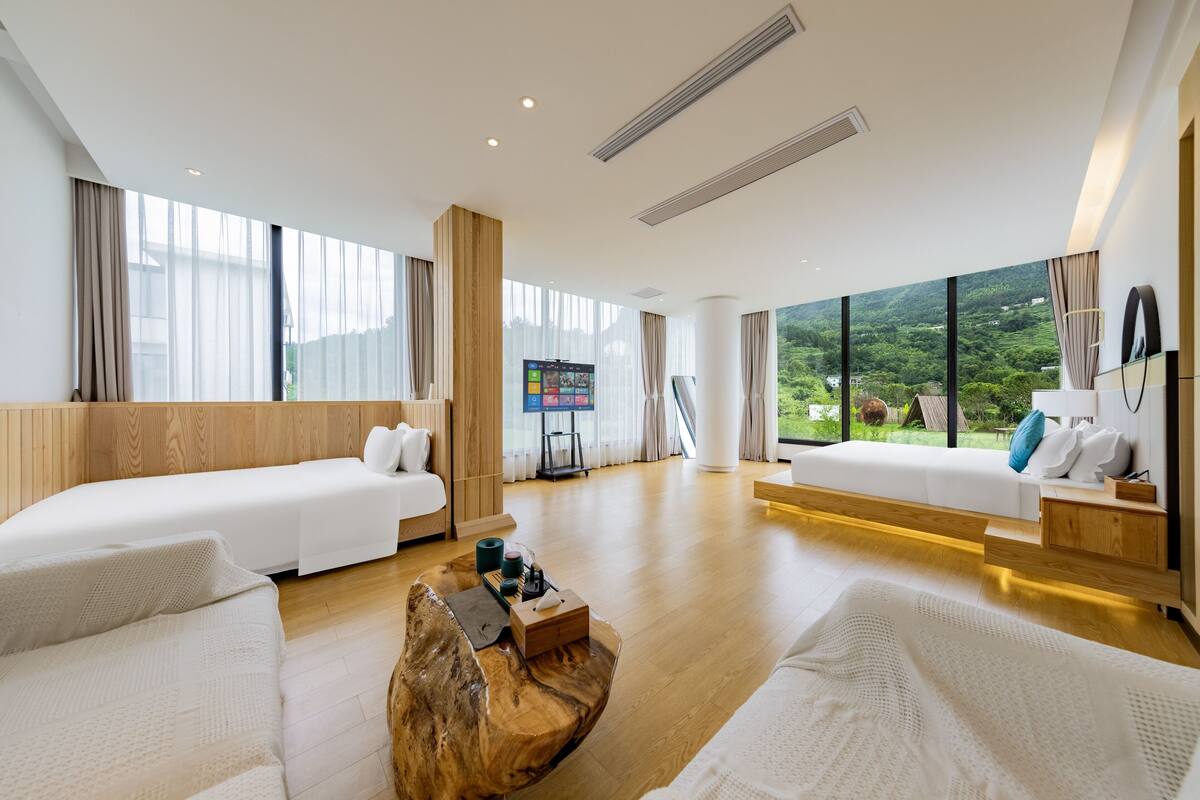 Une chambre d’hôtel moderne avec deux lits, une table basse en bois et une grande fenêtre donnant sur un paysage verdoyant.