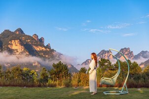 A person standing in a grassy area with a chair and a hanging seat, surrounded by mountains and vegetation.