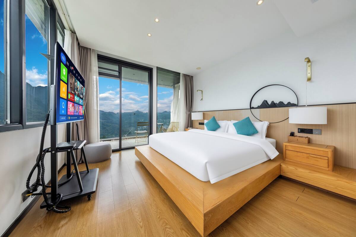 Une chambre d’hôtel moderne avec un grand lit, un téléviseur posé sur un meuble, une table de chevet en bois et une vue sur les montagnes par la fenêtre.
