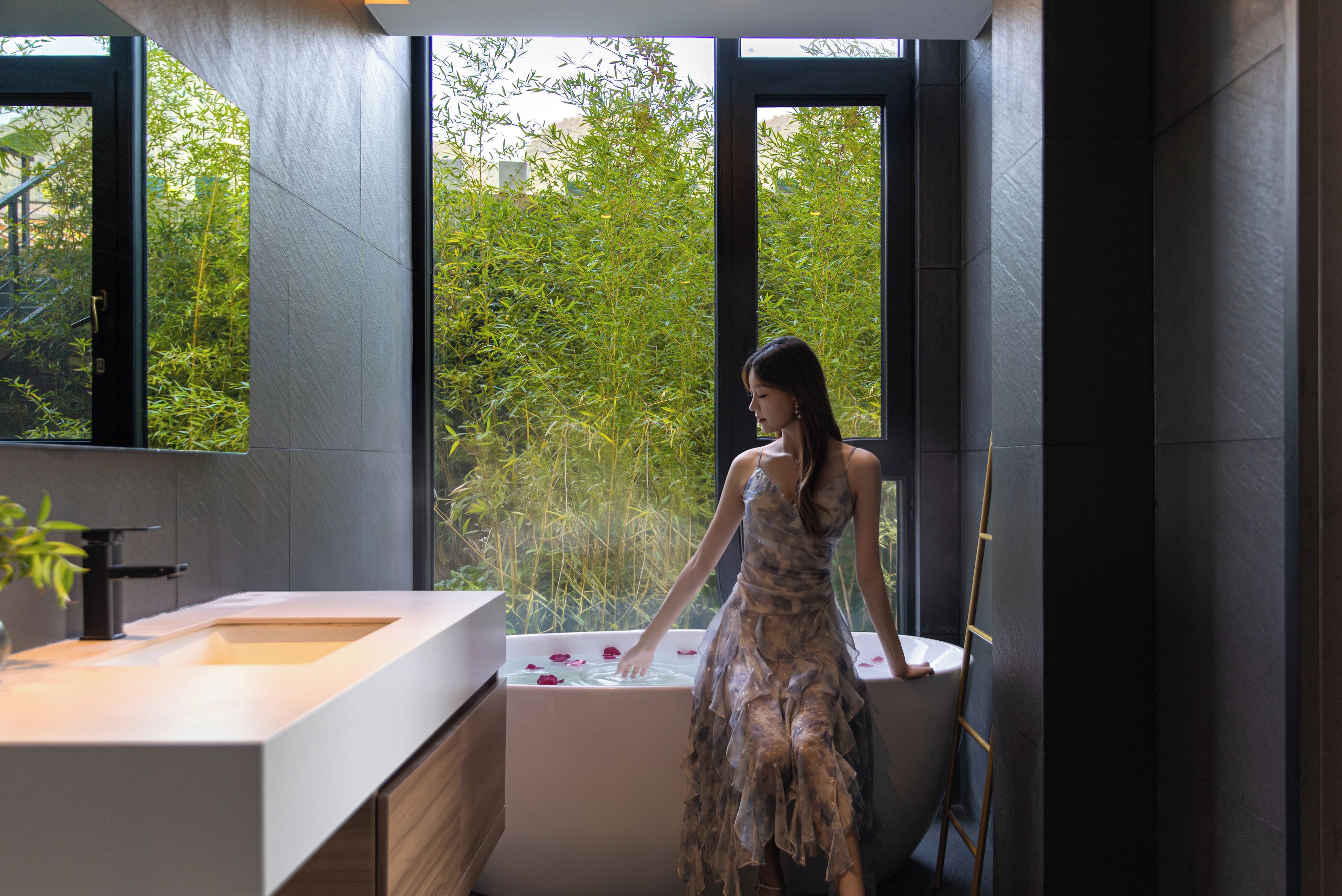 A woman in a dress standing by a bathtub with rose petals, in a modern bathroom with large windows and a view of greenery.
