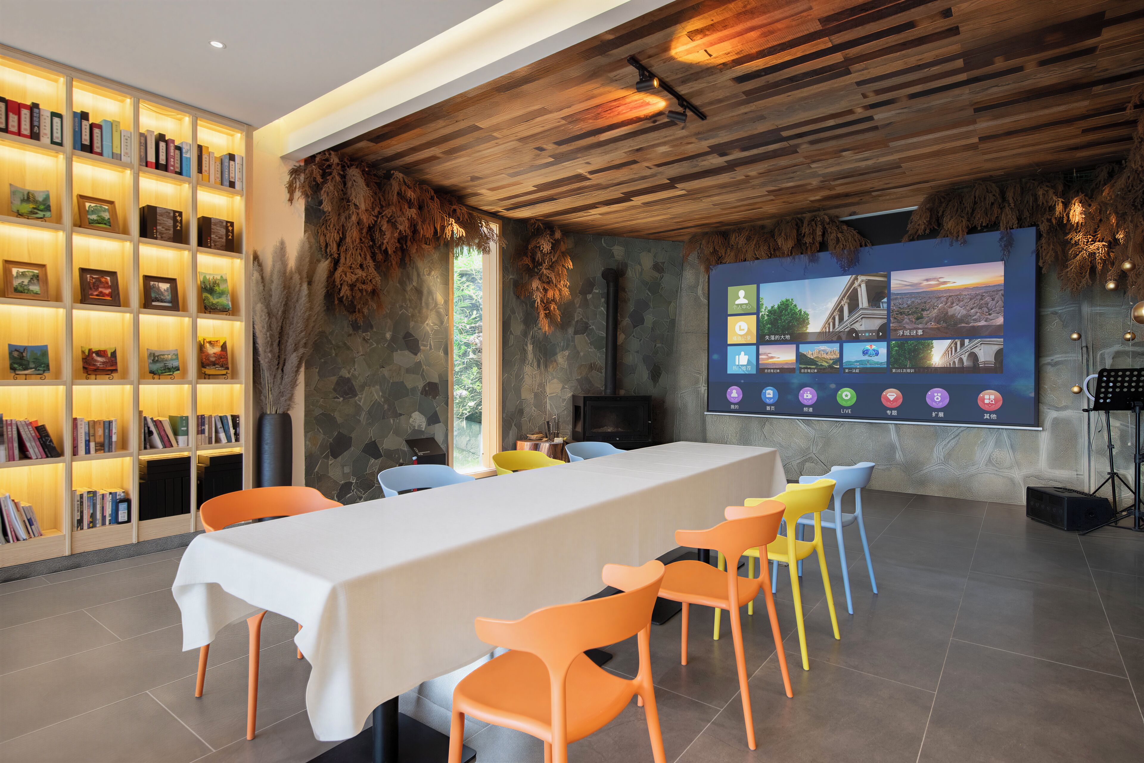 A conference room with a long table, colorful chairs, a bookshelf, and a large screen displaying various images.