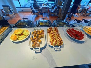 Um buffet com diversos alimentos, incluindo doces e frutas, numa área de refeições com cadeiras e mesas.