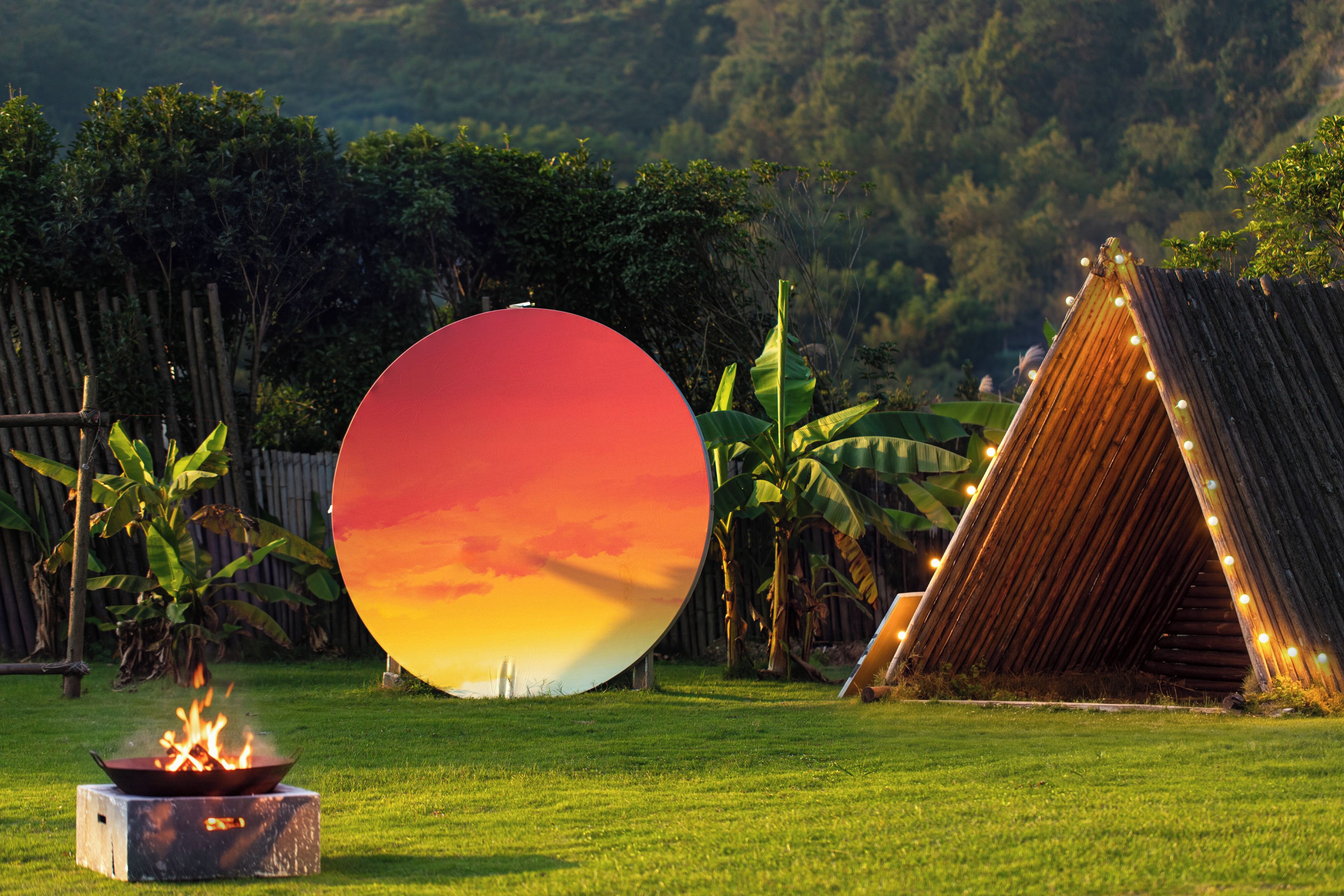 A grassy outdoor area with a fire pit, a large circular object, and a wooden structure with lights.