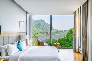 Un dormitorio con una cama grande, una silla, una mesita y vistas a un paisaje montañoso a través de un ventanal amplio.