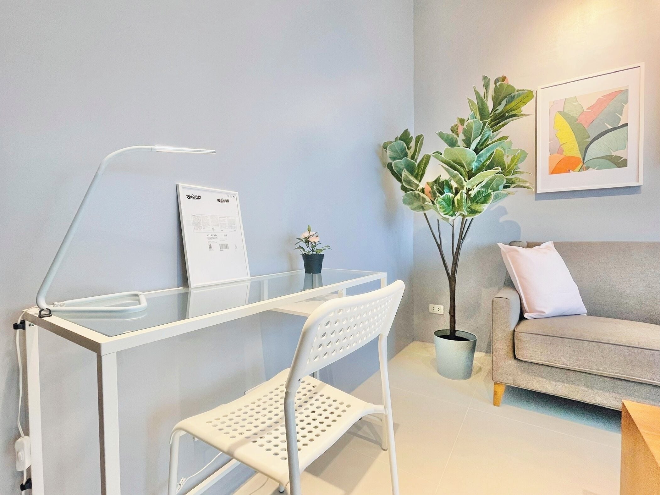 Ein modernes Wohnzimmer mit einem Glastisch, einem weißen Stuhl, einem grauen Sofa, einem Blumentopf und einem gerahmten Bild an der Wand.