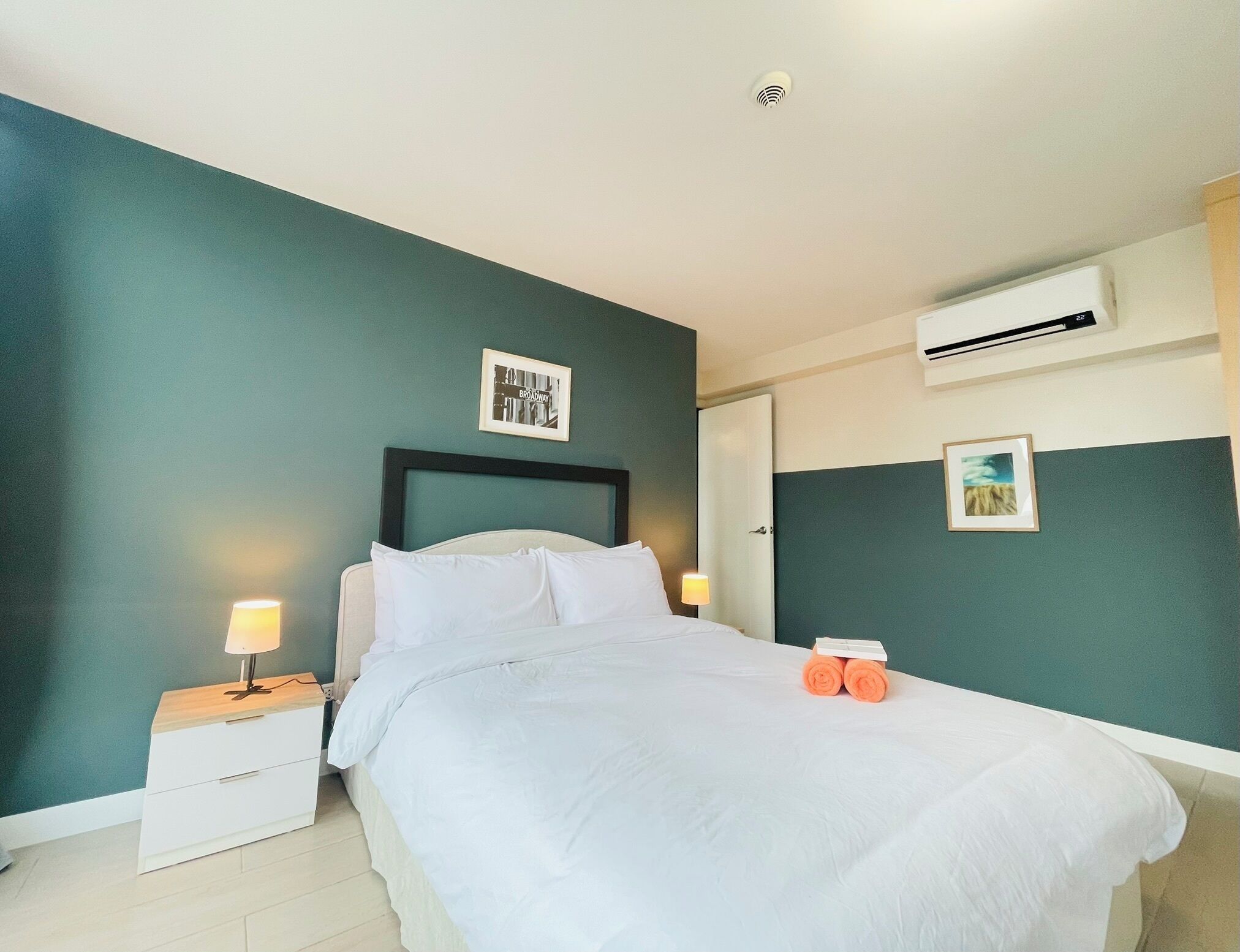 A neatly made bed in a hotel room with teal walls, a white headboard, and two bedside tables with lamps.