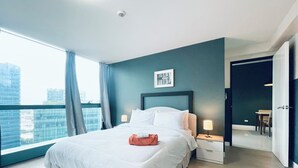 A modern hotel room with a large bed, a nightstand, a lamp, and a view of the city through a large window. - One Uptown Residence Near St Lukes (Taguig)