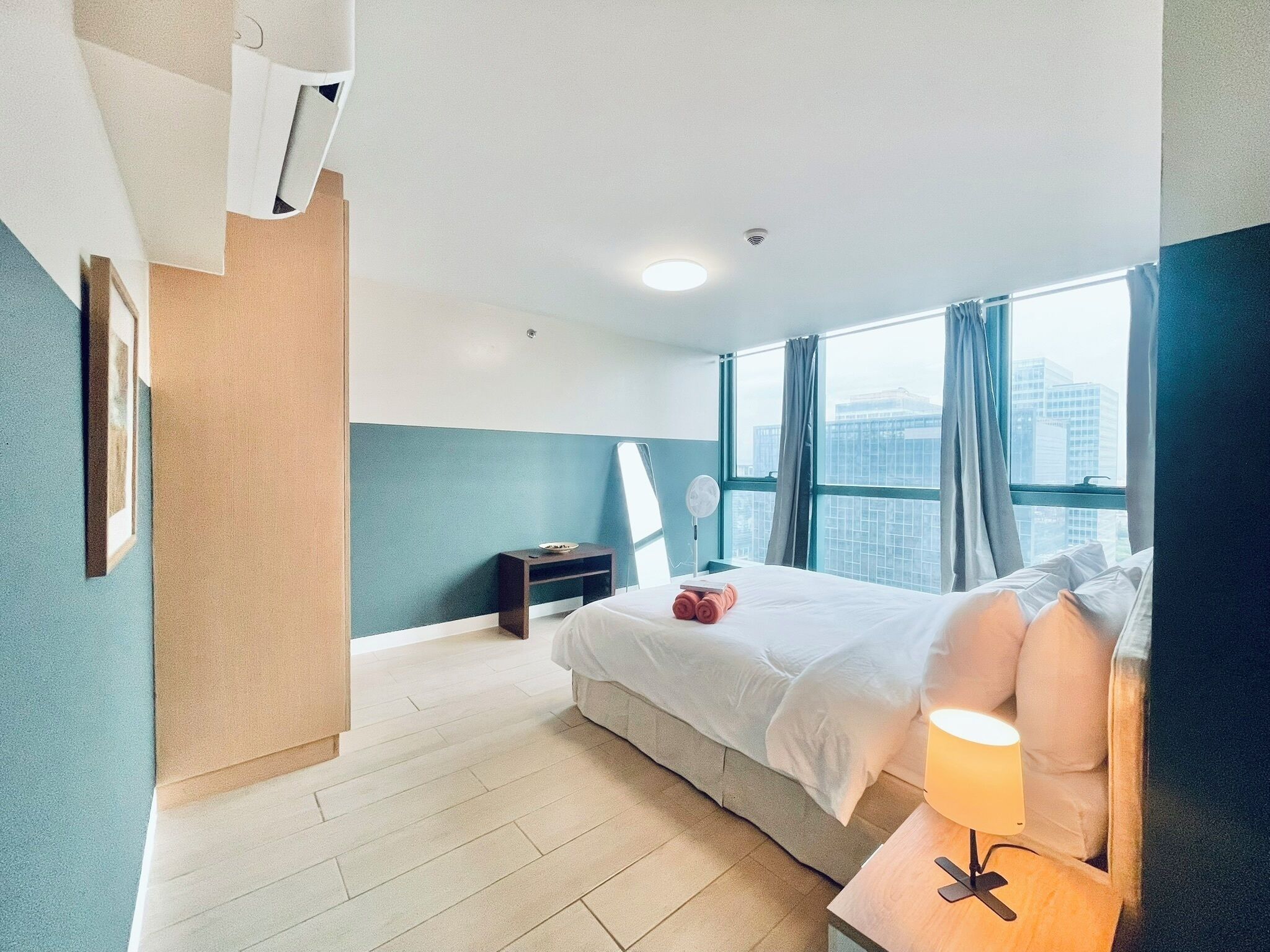 A modern hotel room with a large bed, a bedside table with a lamp, a small nightstand, and a view of the city through large windows.