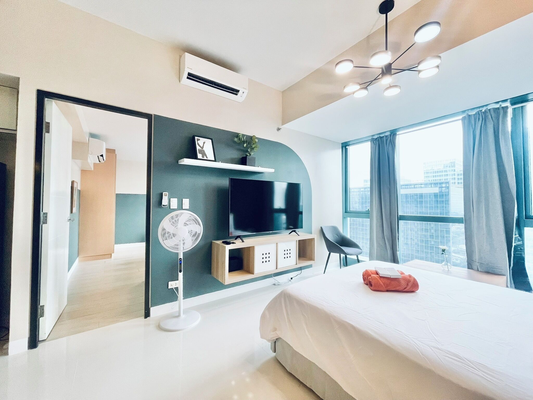 A modern hotel room with a large bed, a flat-screen TV, a standing fan, and a city view through the window.
