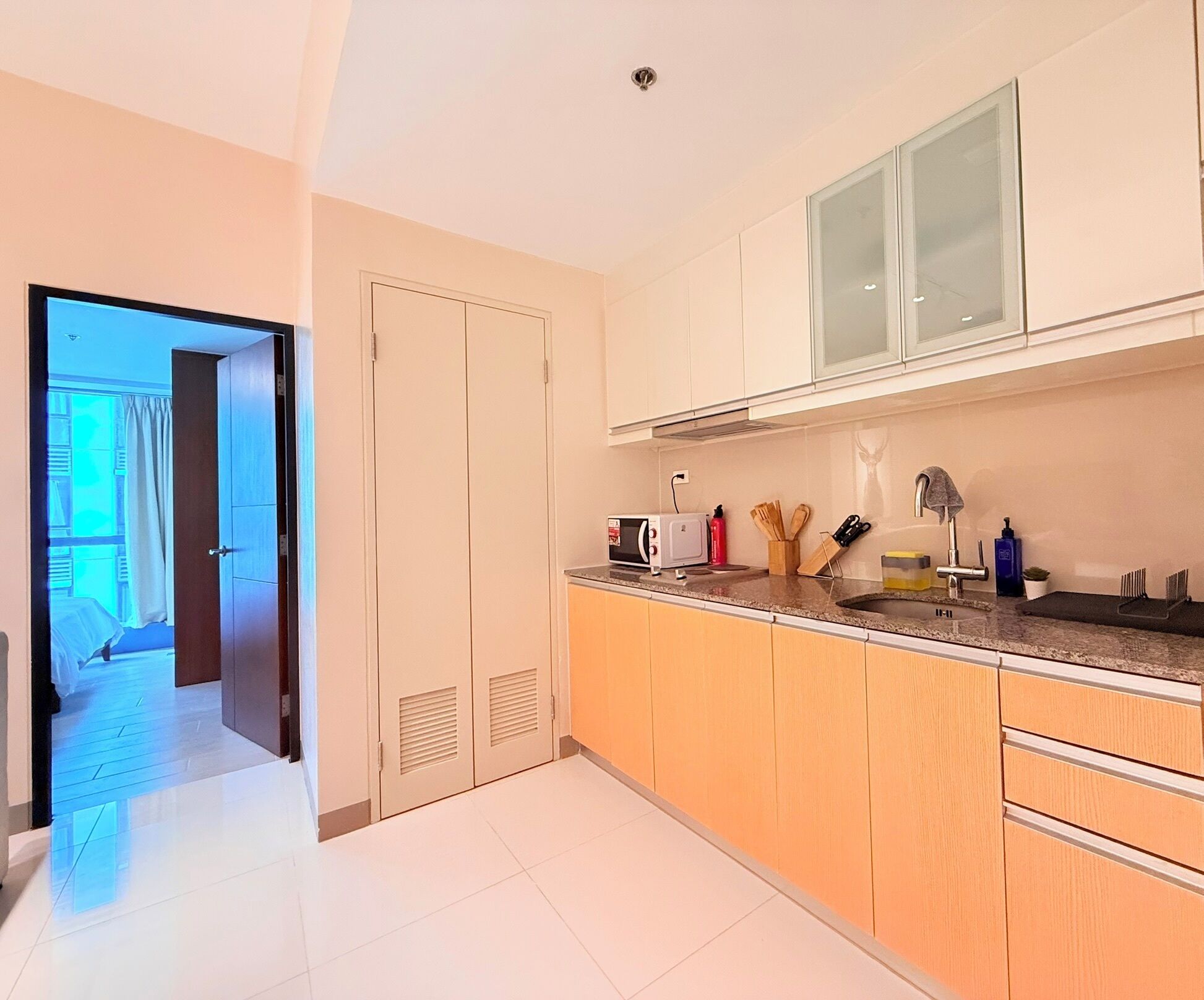 Cozinha moderna com micro-ondas, torradeira e pia. Há uma porta que dá para um quarto com cama e cortinas.