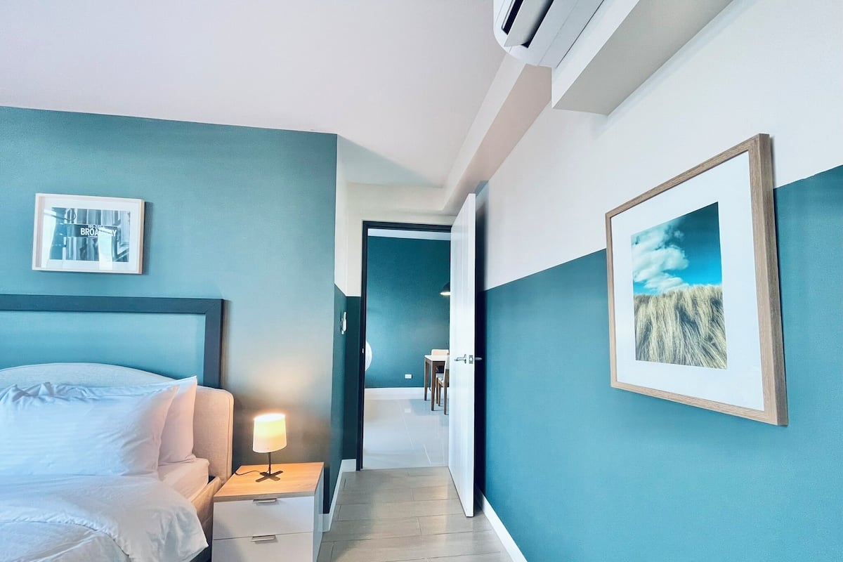 A hotel room with a bed, a bedside table with a lamp, a framed picture on the wall, and a hallway leading to another room.