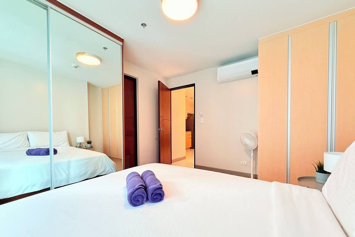 A hotel room with a large bed, a bedside table, a mirror, a wardrobe, and an air conditioning unit.