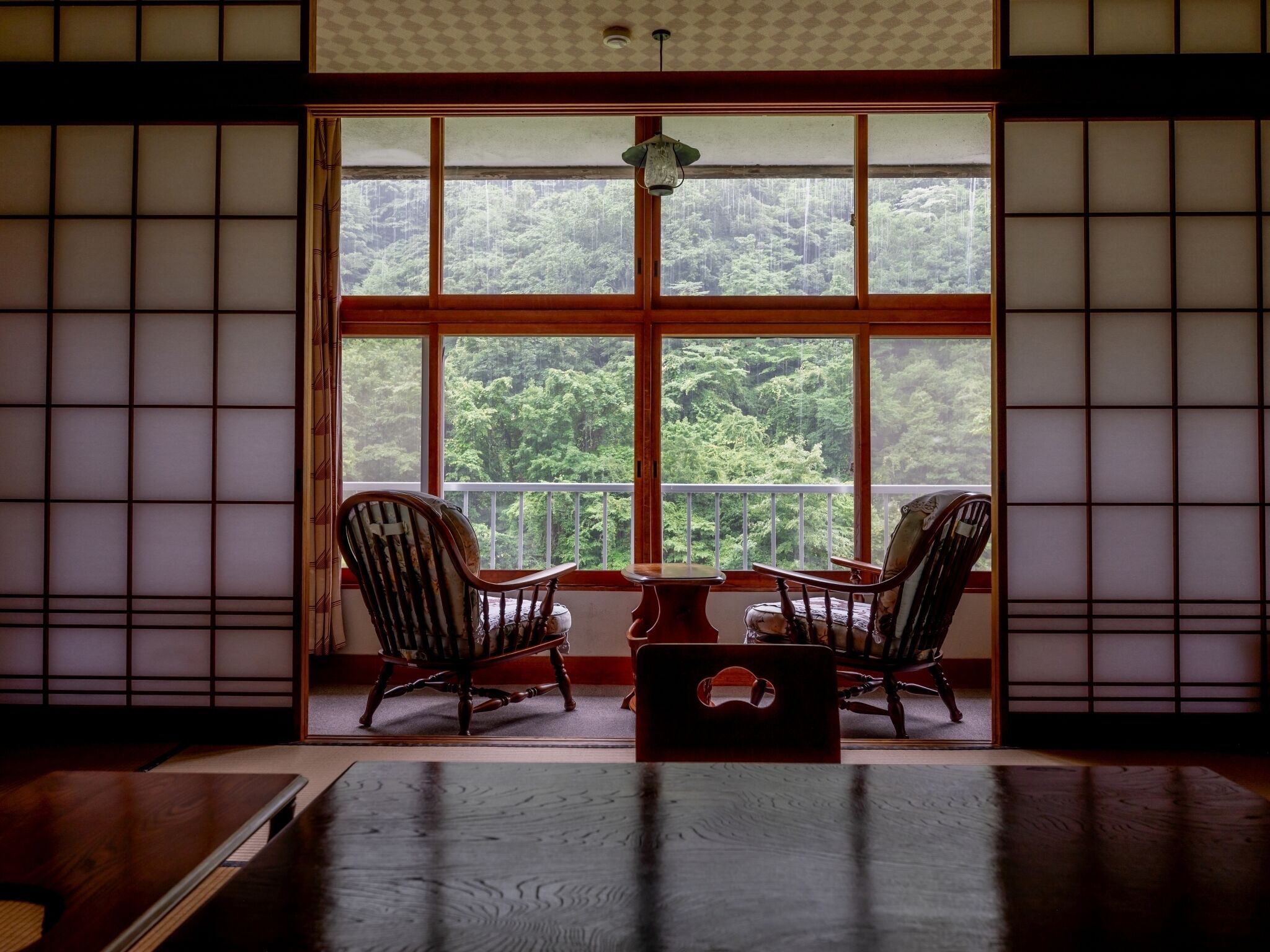 A traditional Japanese room with tatami flooring, a low table, two chairs, and a large window with a view of a forest.