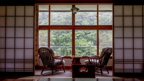 A traditional Japanese room with tatami flooring, a low table, two chairs, and a large window with a view of a forest. - Goshikionsen Goshikinoyu ryokan (Takayama)