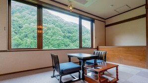 A room with a large window offering a view of a forested hillside. There is a wooden table with chairs and a small wooden cabinet. - Goshikionsen Goshikinoyu ryokan (Takayama)