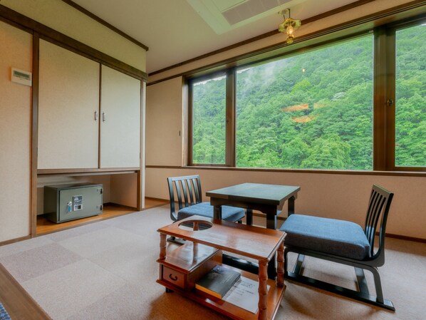 Una habitación con una decoración tradicional japonesa, que incluye suelo de tatami, una mesa baja con cojín y vistas a un bosque frondoso a través de amplias ventanas.