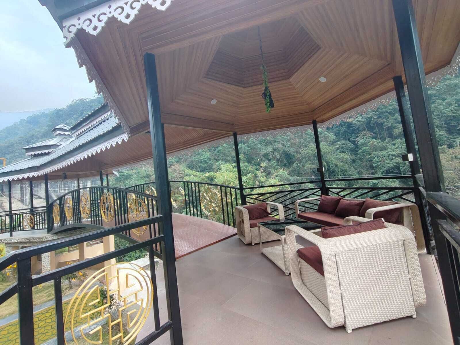 A covered outdoor seating area with wicker furniture, a view of traditional architecture, and a lush forest backdrop.