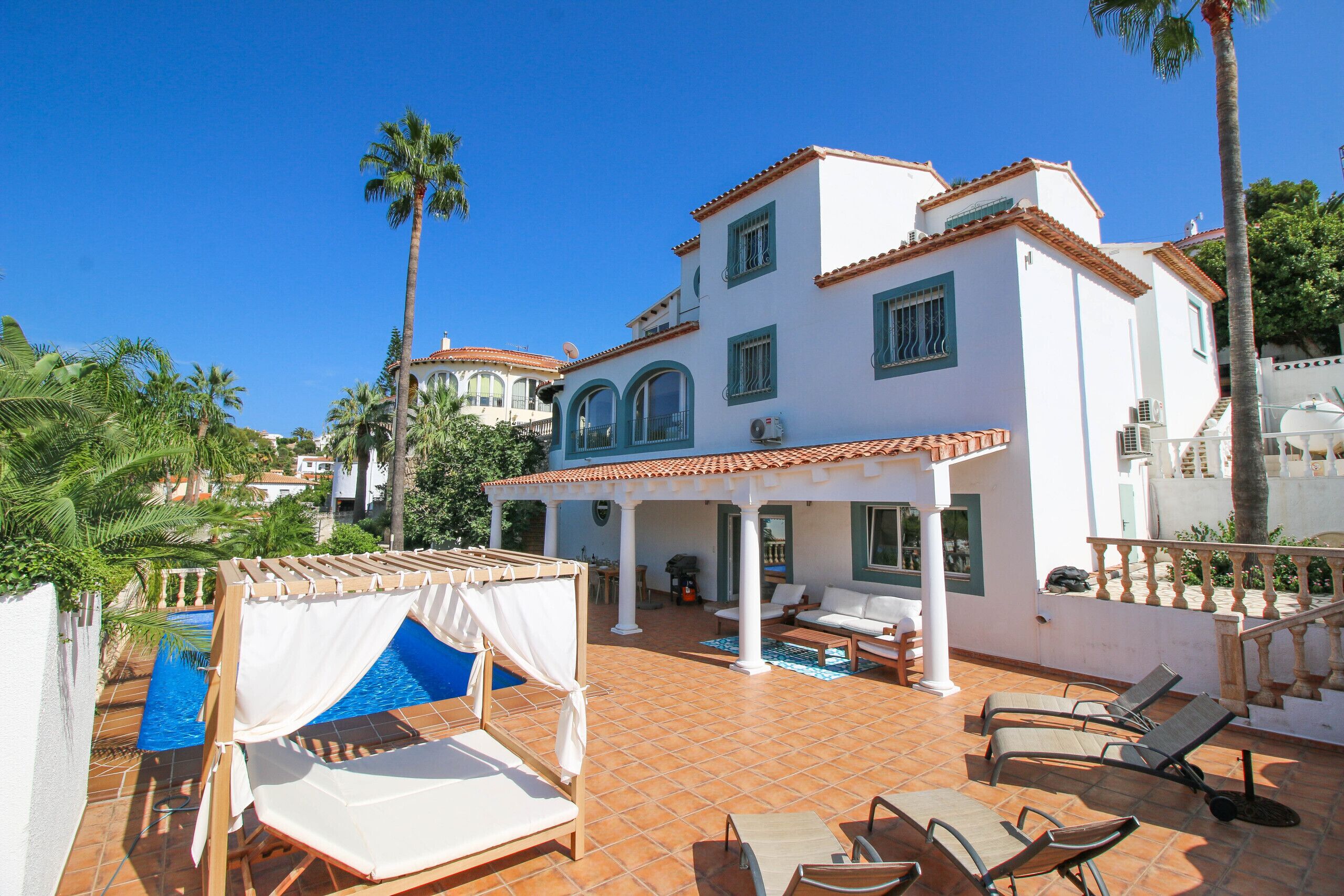 A white villa with a tiled patio, a pool, and lounge chairs.
