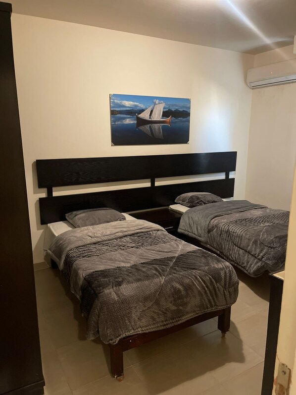 Une chambre avec deux lits, une tête de lit sombre, un tableau représentant un bateau sur l’eau et un appareil de climatisation fixé au mur.