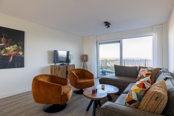 Ein modernes Wohnzimmer mit einer Couch, zwei Sesseln, einem Couchtisch und einem Flachbildfernseher. Ein großes Fenster bietet Blick ins Freie.