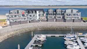 A waterfront property with modern architecture, a marina with boats, and a view of the sea and wind turbines in the background.