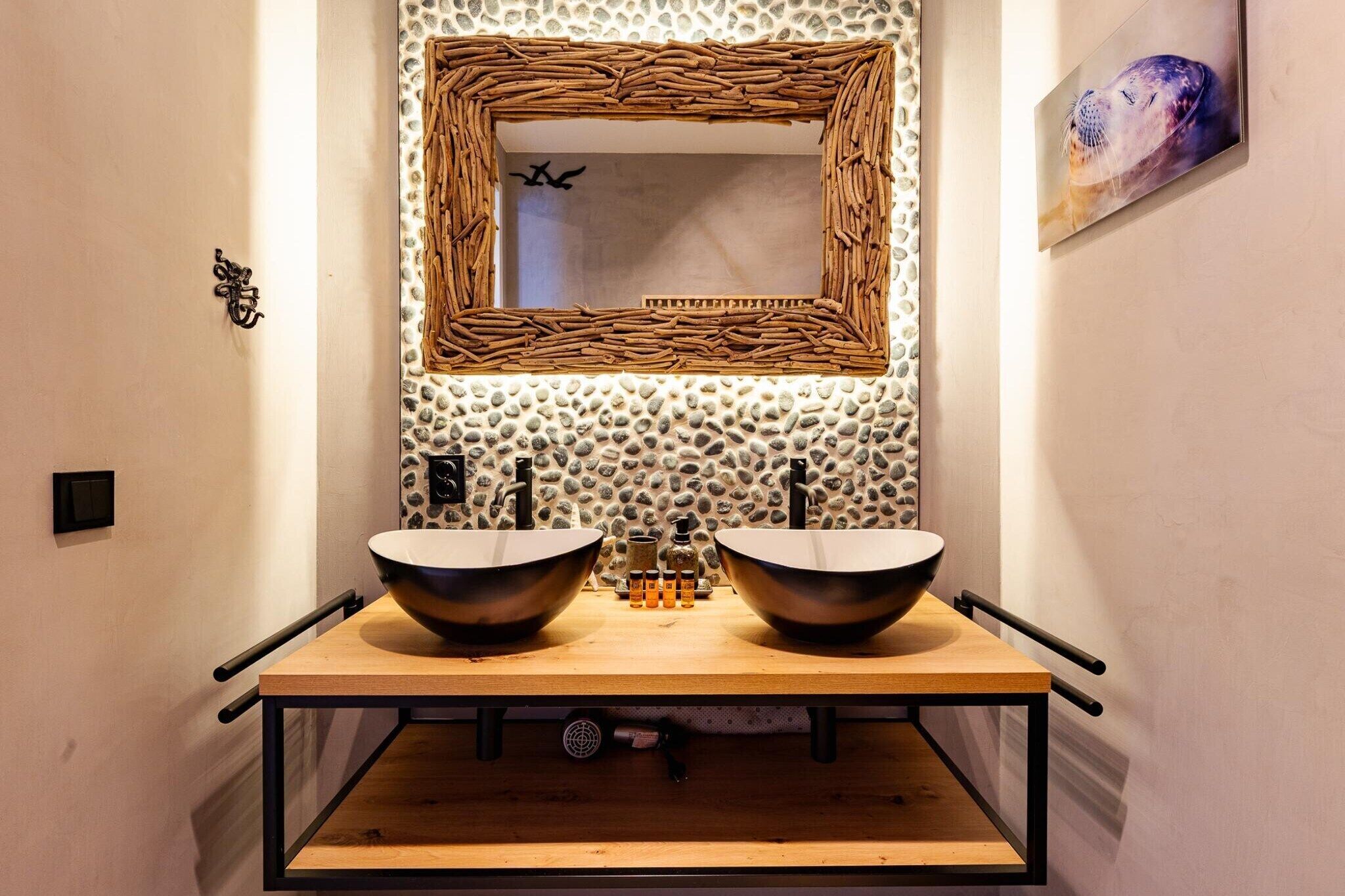 A bathroom with a wooden vanity, two sinks, a mirror with a woven frame, and a stone wall.