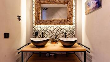 A bathroom with a wooden vanity, two sinks, a mirror with a woven frame, and a stone wall.