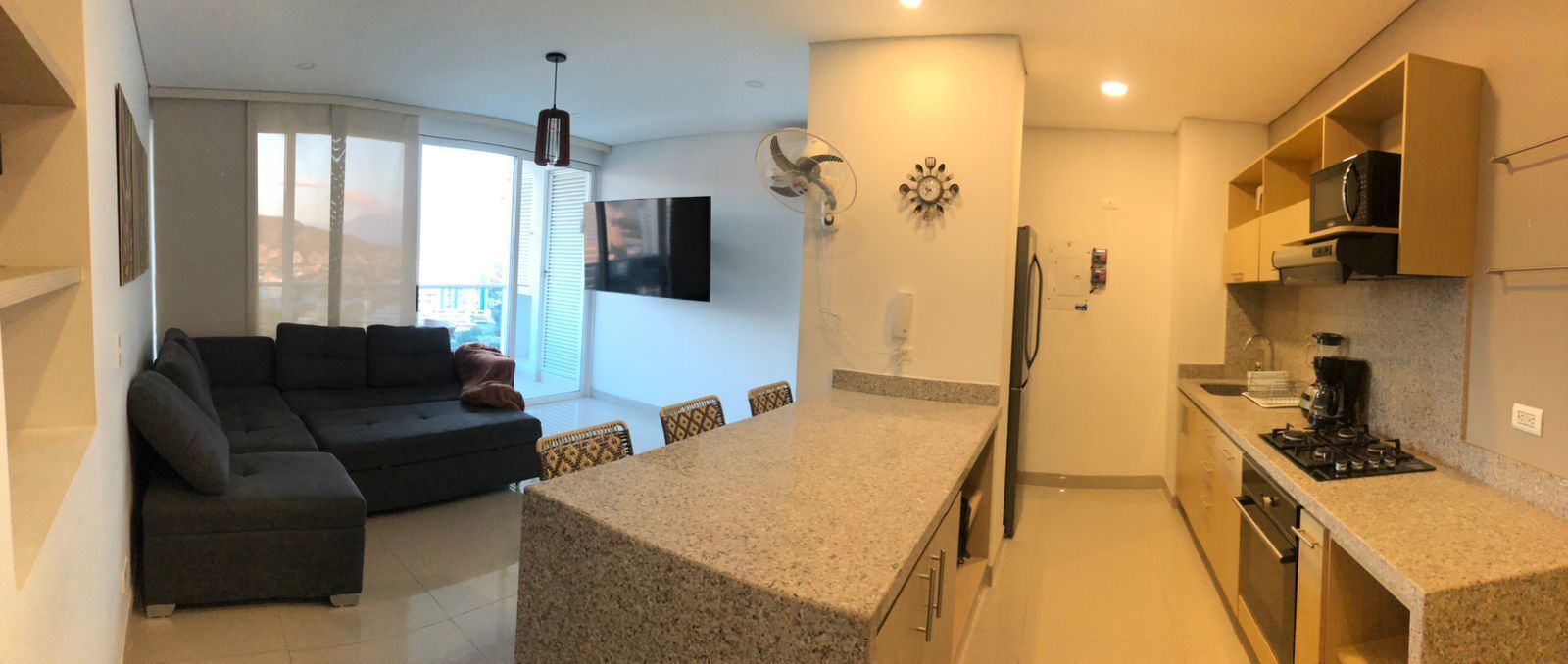 A compact living space with a sofa, dining table, kitchen area, and a TV mounted on the wall.