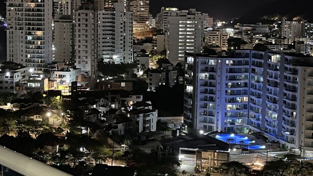 Vista nocturna de un paisaje urbano con rascacielos, una zona de piscina con iluminación azul y montañas al fondo.