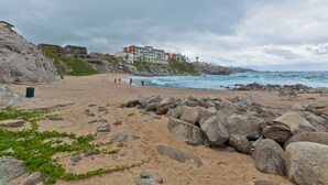A beach with large rocks, a few people, and a building with multiple balconies overlooking the ocean. - Casa Famlee - Duplex House(sleeps 8) with Pool & Hot Tub in Cabo San Lucas! (Cabo Bello)