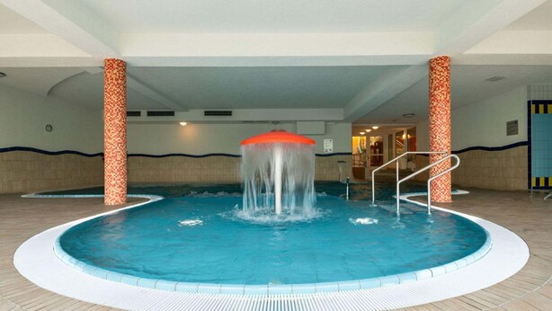 Una piscina interior con un elemento acuático central, rodeada de suelo de baldosas y sostenida por columnas con bases decoradas.