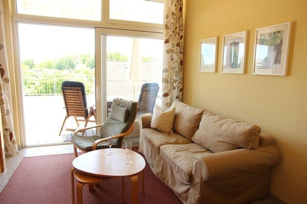 A living room with a beige sofa, two chairs, a round table, and a glass door leading to a balcony.