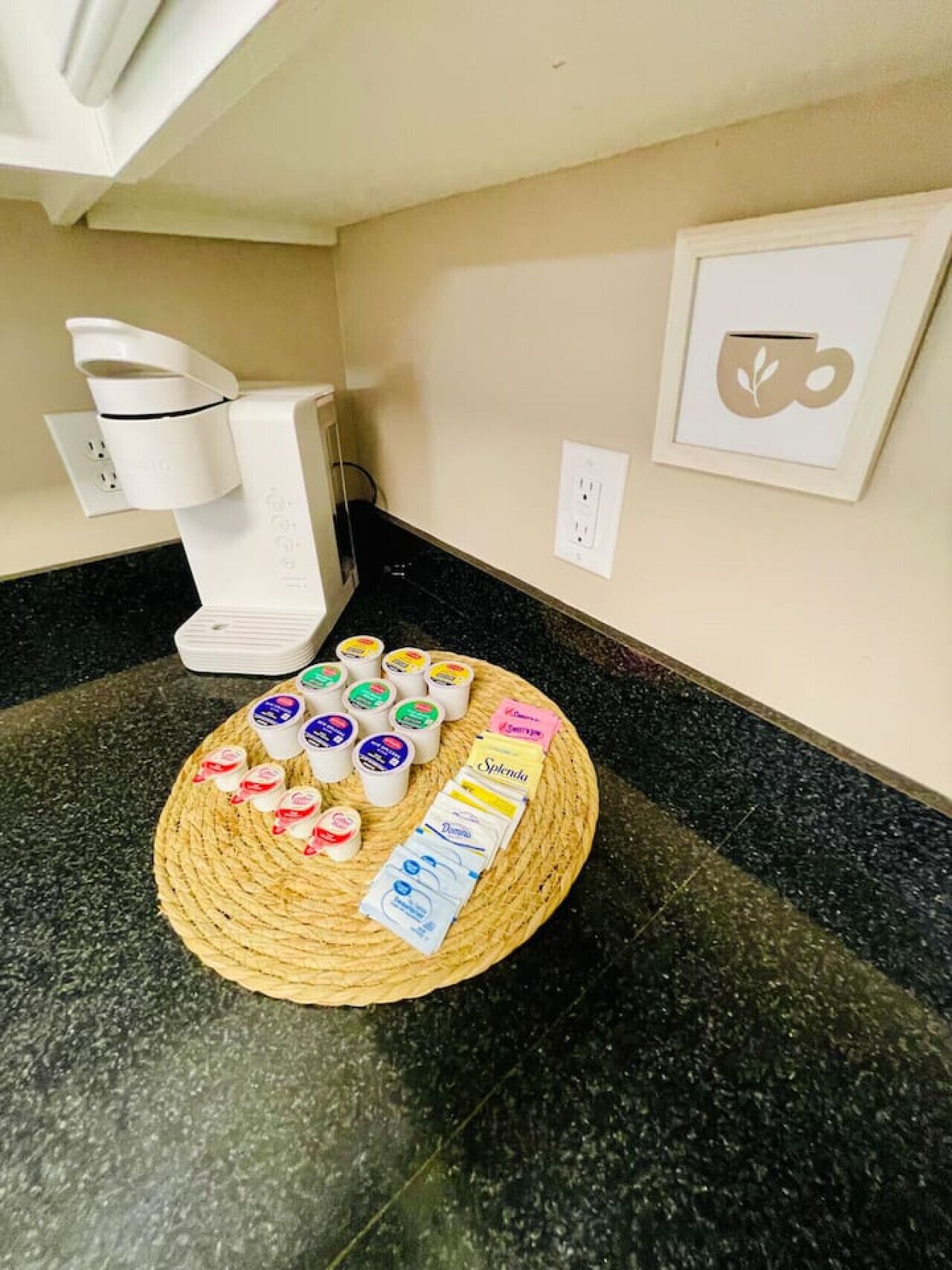A kitchen counter with a coffee maker, a woven mat with packaged food items, and a framed picture on the wall.