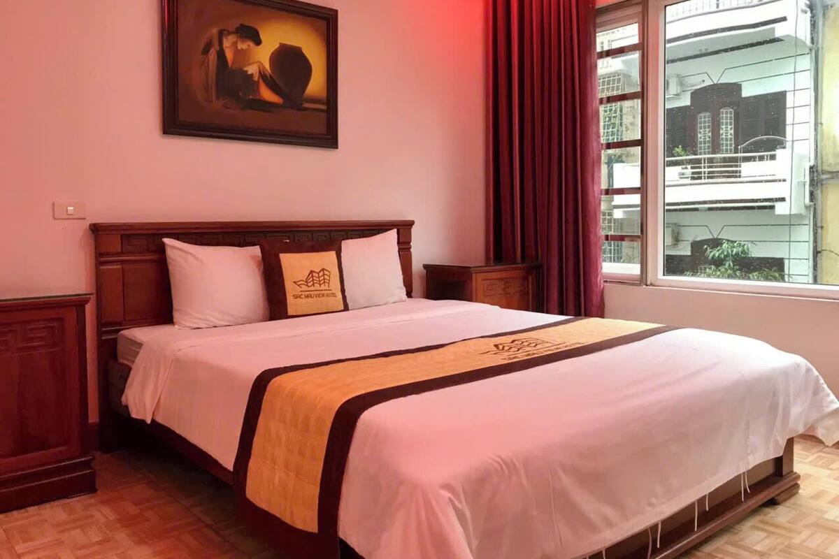 A hotel room with a large bed, a wooden headboard, a painting on the wall, and a window with curtains.