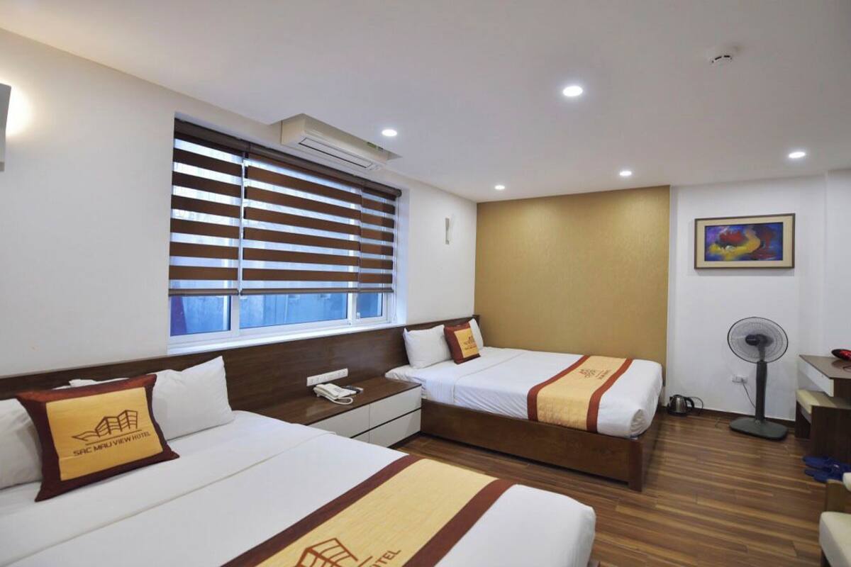 A hotel room with two beds, a bedside table with a phone, a standing fan, and a window with blinds.