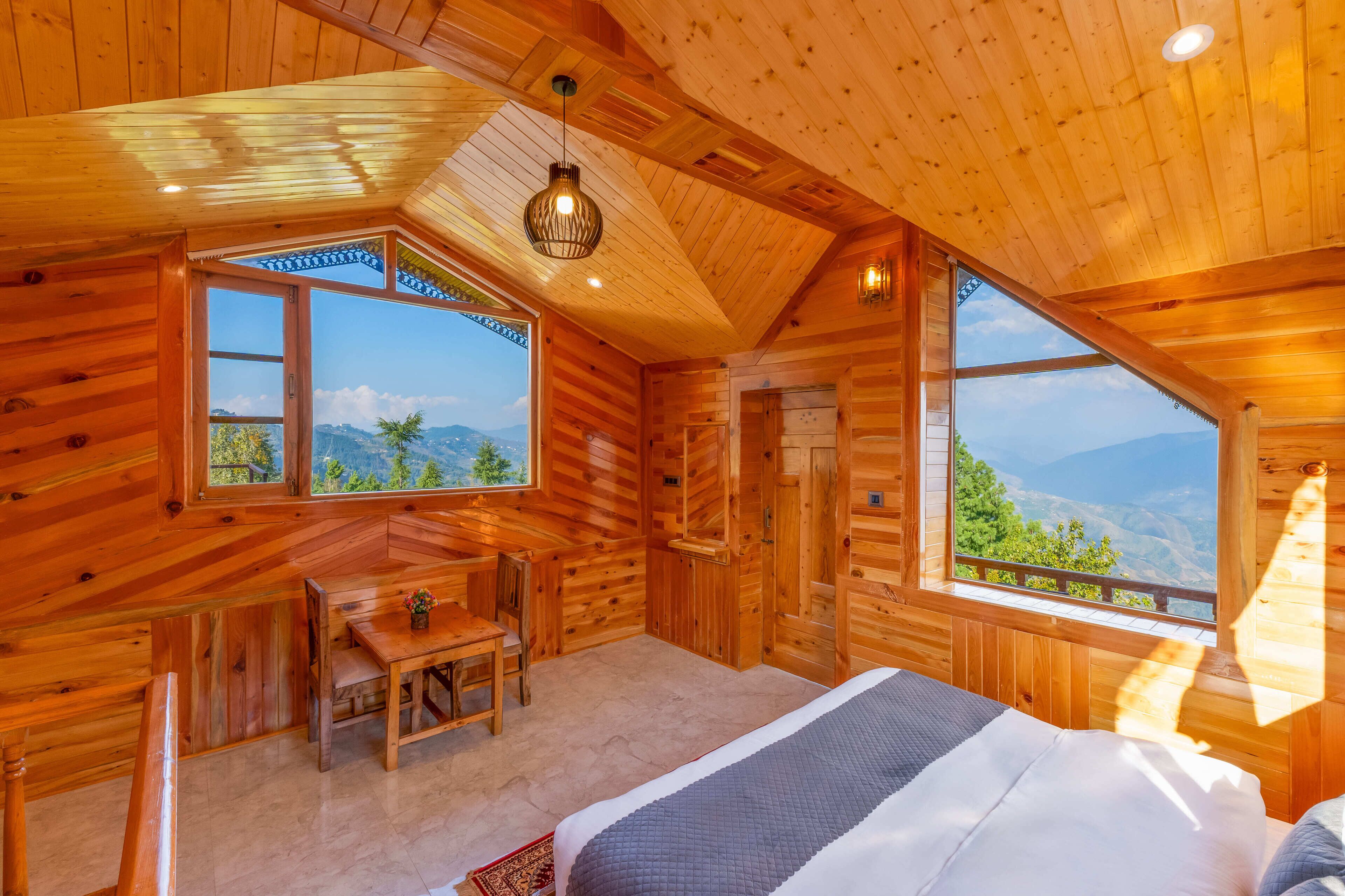 Camera da letto in una baita di legno con un letto, una scrivania e una sedia. Ampie finestre offrono vista sull'esterno.