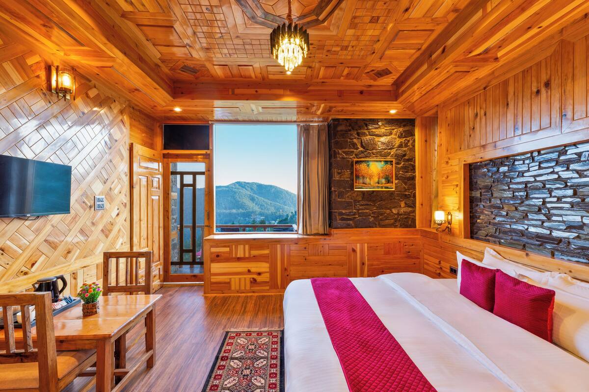 Interior de una cabaña de madera con una cama, un televisor, un muro de piedra y vistas a las montañas.
