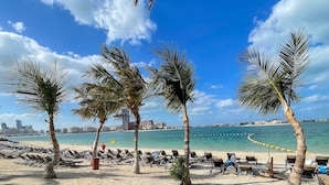 A beach with sun loungers, palm trees, and a city skyline in the background. - LUX Dream Palm Atlantis Sea View Suite (Dubai)