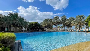 A large, clear swimming pool surrounded by palm trees and lounge chairs, with a view of a distant cityscape. - LUX Dream Palm Atlantis Sea View Suite (Dubai)