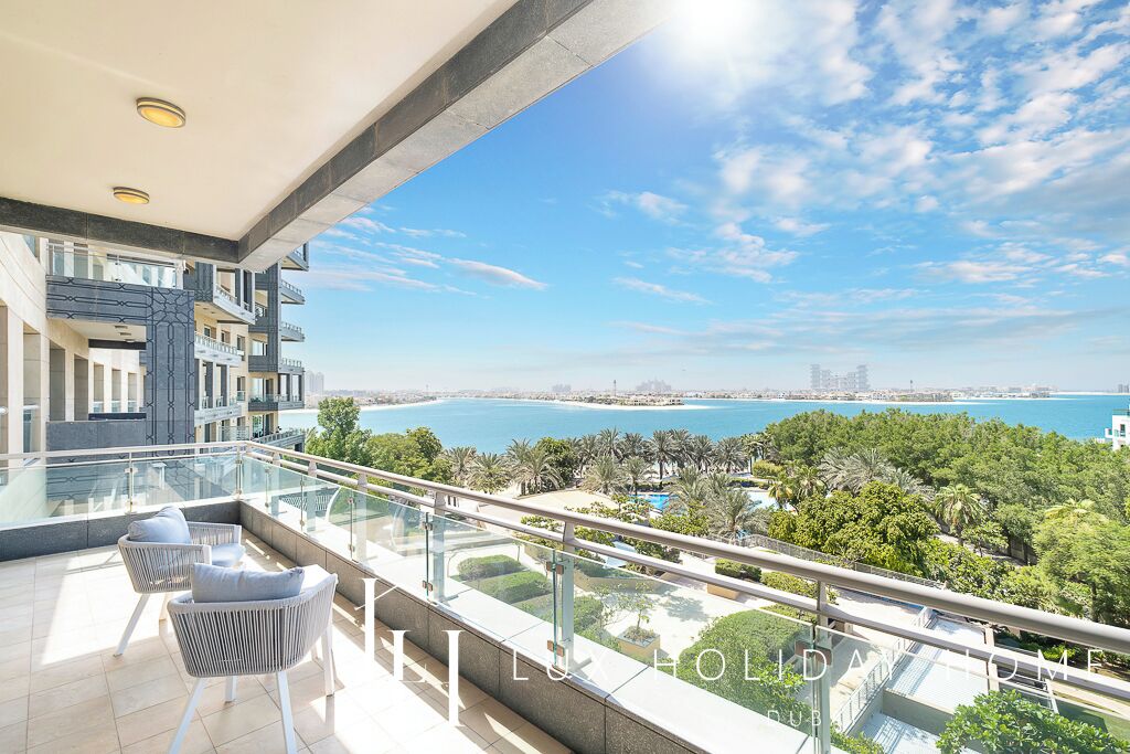 A balcony with a glass railing overlooking a coastal view, a pool, and a cityscape in the distance.