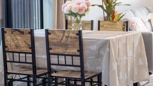 Un coin repas avec une table dressée pour quatre personnes, entourée de chaises en bois dont les pieds sont en métal. Un vase rempli de fleurs roses est posé sur la table, et une grande plante en pot se trouve à l’arrière-plan.