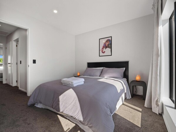 A neatly made bed with a grey and white duvet, a framed picture on the wall, and a bedside table with a salt lamp.