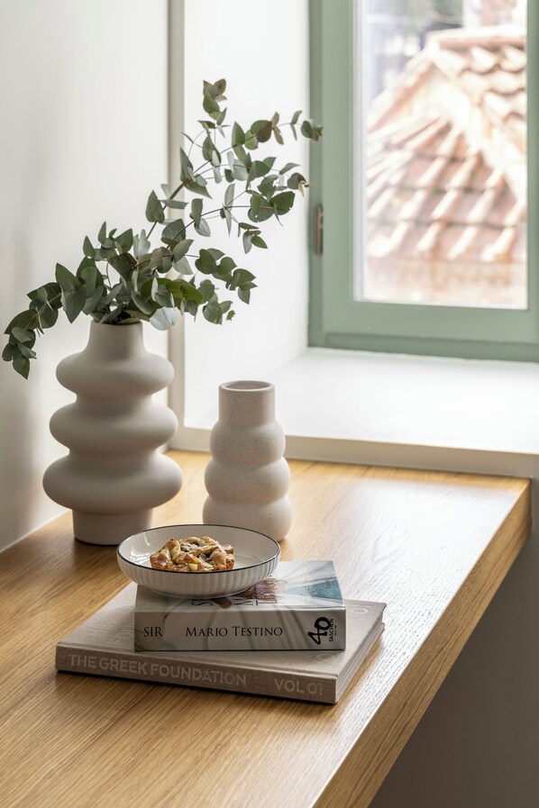 A wooden table with a glass bowl of nuts, two books, and a white vase with green leaves. - Whisper Athens Suites. (Athens)