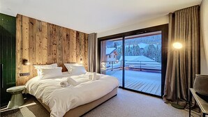 Un dormitorio moderno con una cama grande, paneles de madera en las paredes, un gran ventanal corredero a un balcón y vistas de un paisaje nevado.
