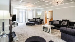 Un salon spacieux avec un plafond haut, un lustre et une cheminée. On y trouve deux canapés avec des coussins à motifs et une table basse au centre.