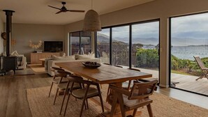 Un vaste espace de vie comprenant une grande table à manger en bois, des sièges confortables, un ventilateur de plafond et une grande fenêtre offrant une vue panoramique sur l’eau et les collines.