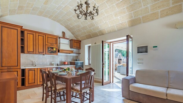 Cozinha espaçosa com armários de madeira, mesa de jantar com cadeiras e um sofá bege em ambiente claro com piso em azulejo e teto de pedra.