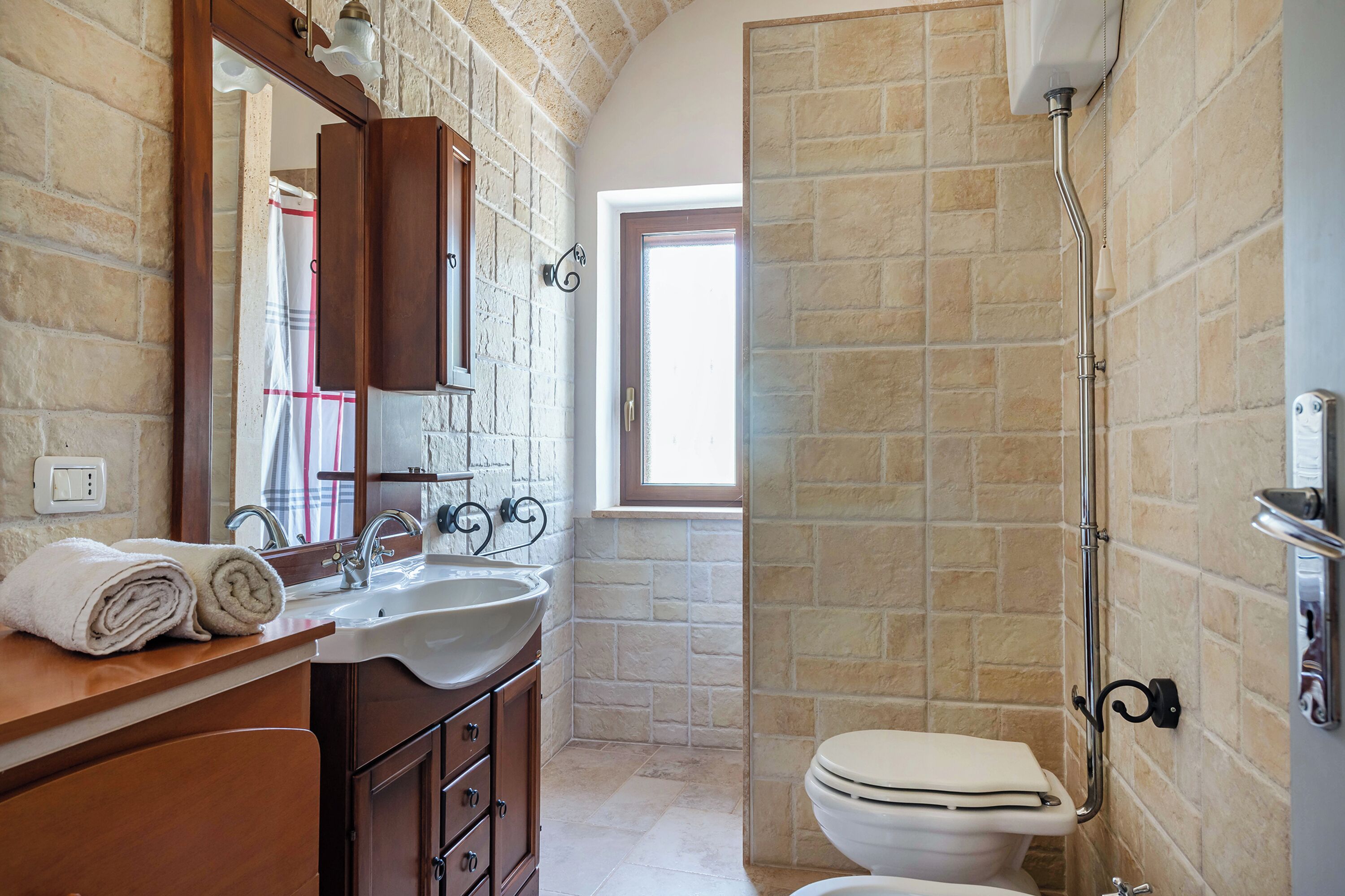 Un bagno con parete in pietra, un lavandino, un water e una finestra che fa entrare luce naturale.