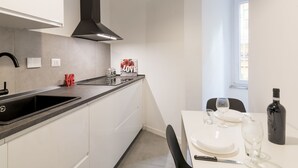 A modern kitchen with white cabinetry, a black countertop, and a black range hood. There is a dining area with a table set for two, a wine bottle, and a glass. - Welcome Rome by Interhome (Rome: Historical City Center)